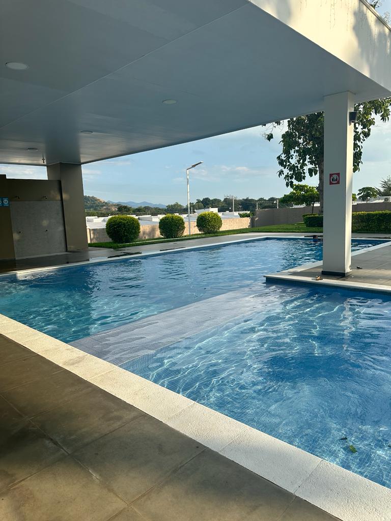 A refreshing resort-style pool is surrounded by greenery, with clear blue water gently rippling under sunlight. The area features a covered space, offering shaded seating options, and is framed by neatly trimmed hedges, providing a serene environment for relaxation.