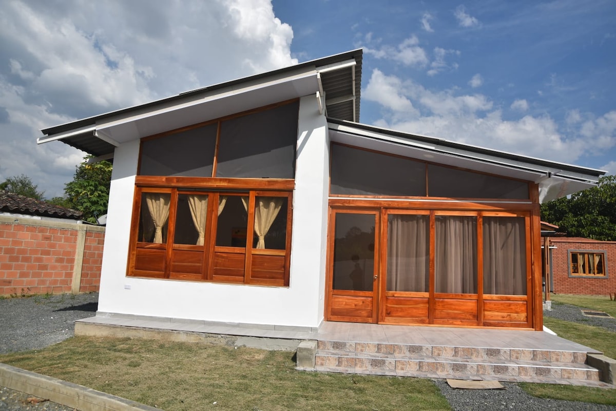 A modern cabin exterior is displayed, featuring large wooden-framed windows. A welcoming entrance is highlighted by stone steps leading up to a covered porch. Light-colored walls contrast with wooden accents, while a spacious lawn surrounds the cabin, inviting outdoor relaxation.