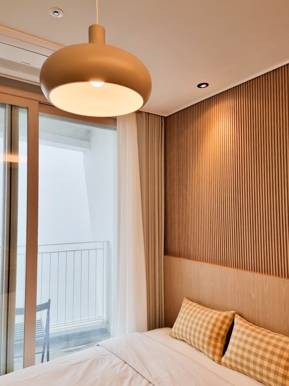 A softly lit bedroom features a queen-sized bed with a white duvet and two checkered pillows. Natural light filters through sheer curtains, illuminating the textured wooden wall behind the bed. A modern pendant light hangs from the ceiling, complementing the cozy ambiance.