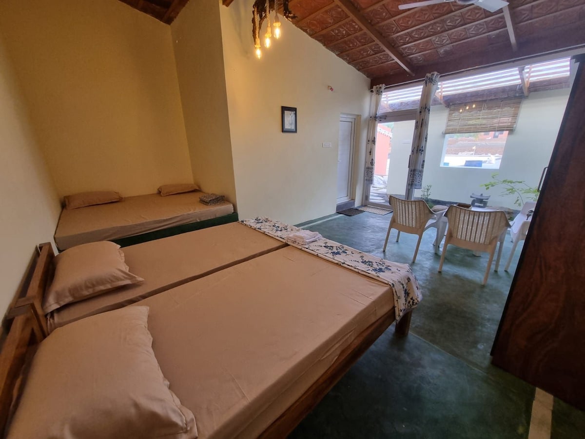 A spacious room features two cozy beds arranged with neutral-toned bedding. Natural light enters through large windows, illuminating the space. A small seating area with two chairs and a table is set beside a potted plant, enhancing the room's relaxing atmosphere.