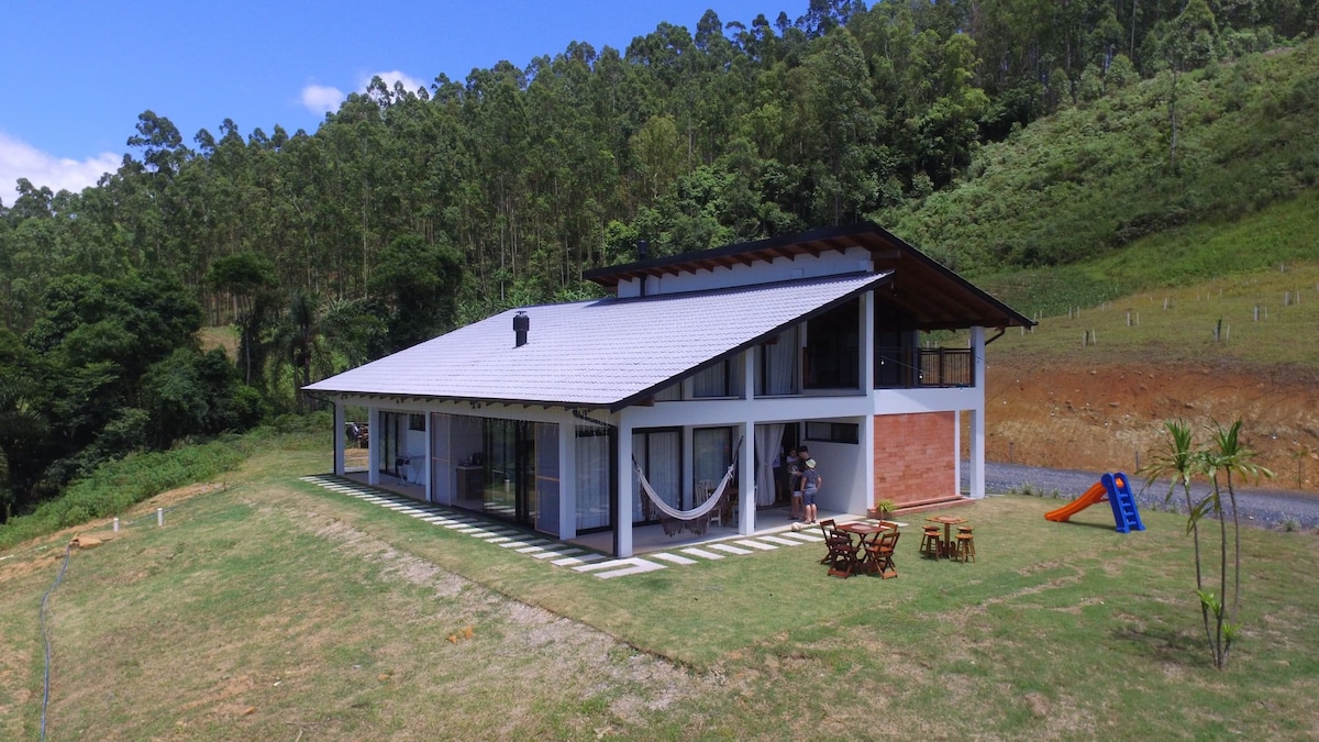 The exterior of the modern house features a spacious design with large glass doors and a sloping roof. A hammock is draped on a covered porch, and outdoor seating is arranged on the lawn, with a colorful slide visible nearby, all set against a backdrop of lush greenery.