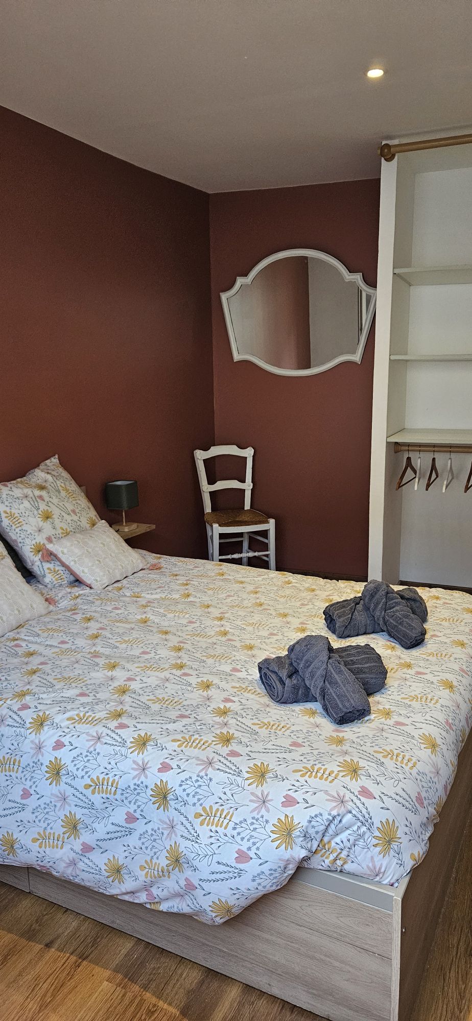 A cozy bedroom features a double bed with a floral-patterned duvet cover. A mirror hangs on the wall above a small nightstand, and a wooden chair is positioned nearby. Light wood flooring complements the warm, earthy tones of the walls.