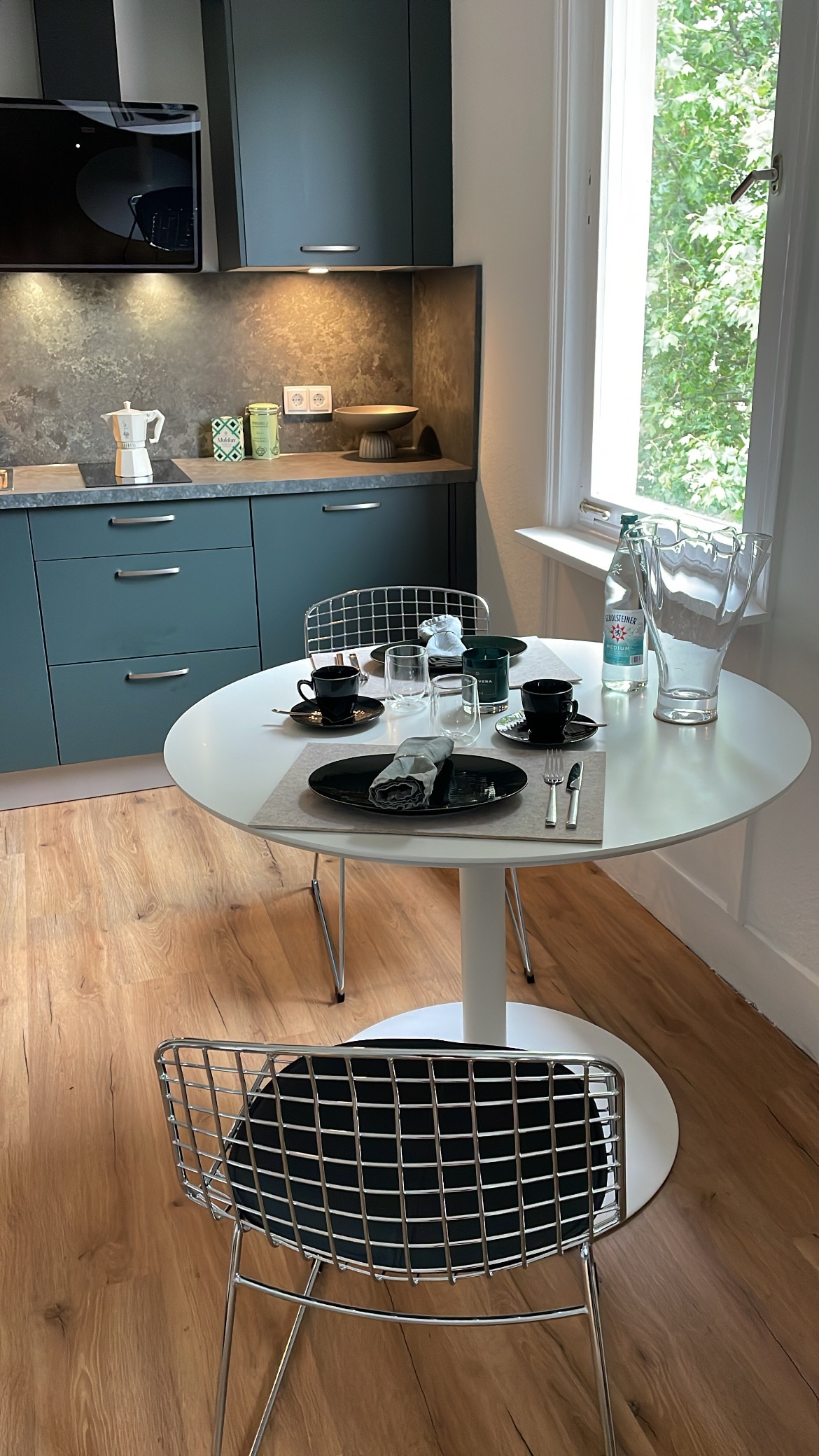 A modern kitchen area features a round white table set for two, with black plates, silver cutlery, and glassware. Wire chairs provide seating, while a backdrop of dark cabinetry and light countertops complements natural light coming through a window with greenery outside.