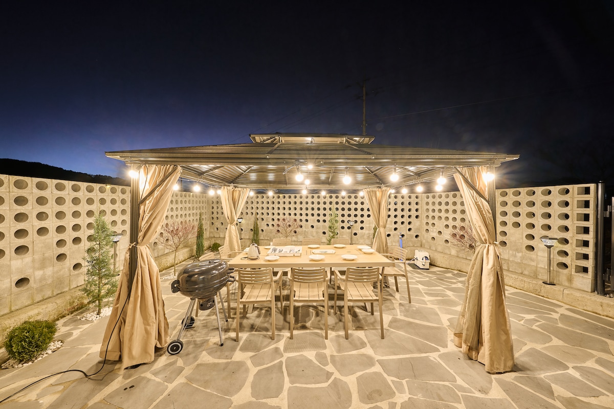 An outdoor barbecue area is showcased under a canopy with soft lighting, creating a welcoming space for gatherings. A large wooden table and multiple chairs are arranged for dining, while a grill is positioned nearby. Surrounding walls feature modern perforated designs.