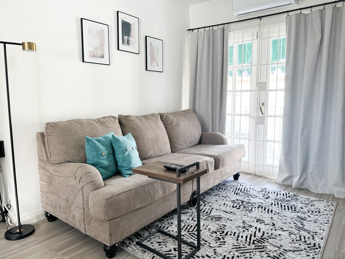 A light-filled living area is shown, featuring a comfortable sofa adorned with teal accent pillows. A small coffee table rests nearby, and sheer curtains frame the glass doors that lead outside. Wall art is displayed above the sofa, enhancing the modern decor.
