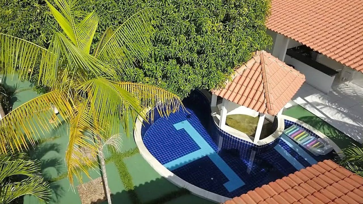 An aerial view showcases a private outdoor area featuring a turquoise-tiled pool surrounded by lush greenery. A gazebo with a tiled roof is positioned nearby, providing shade. Brightly colored lounge chairs are arranged along the poolside, enhancing the inviting ambiance of the space.