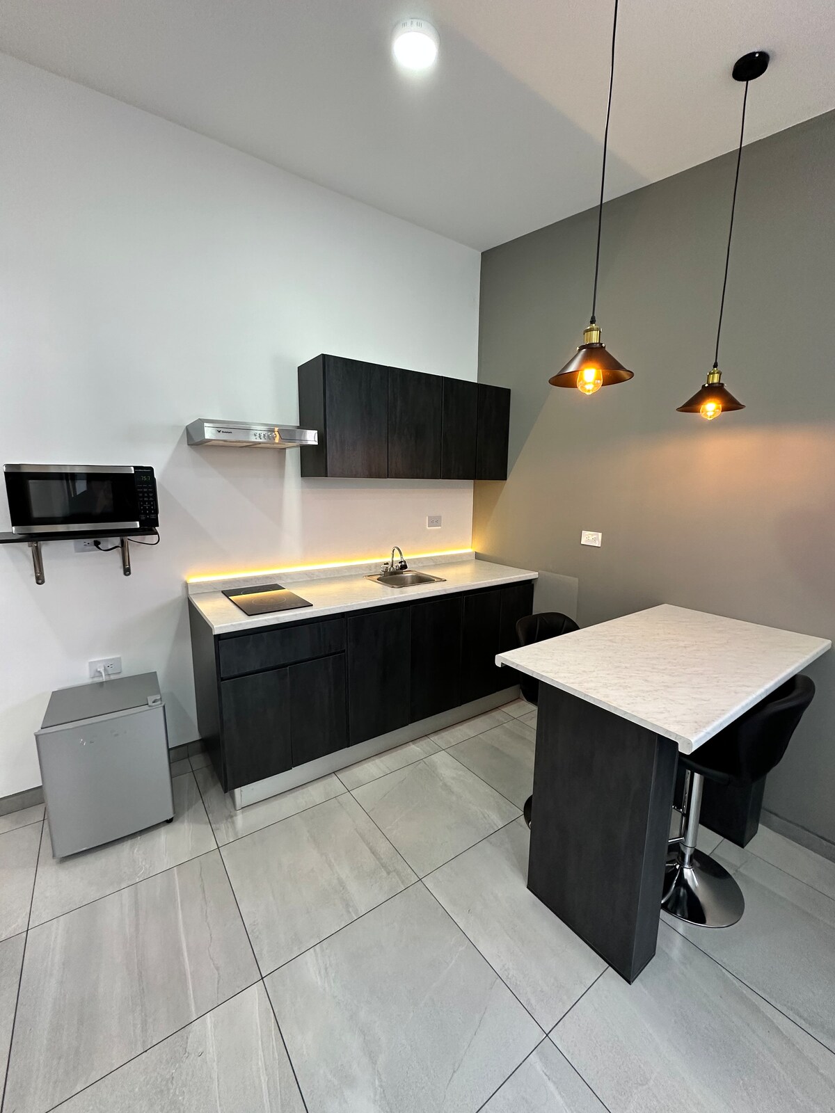 A modern kitchen features dark cabinetry and a countertop illuminated by warm lighting. A stainless-steel sink is positioned beneath a shelf, complemented by a small microwave and a compact refrigerator. Two stylish bar stools are placed at a high table, enhancing the kitchen's functionality.