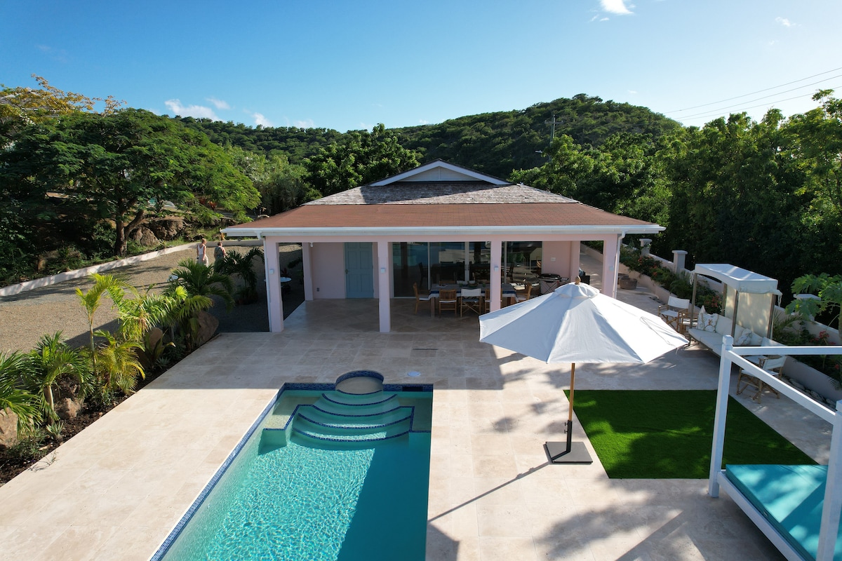 An aerial view captures the Pink House, highlighting the private infinity pool with a spa feature. The spacious outdoor terrace includes a sun umbrella and lounge area, surrounded by tropical greenery. Ample seating is visible, offering inviting space for relaxation and enjoying views.