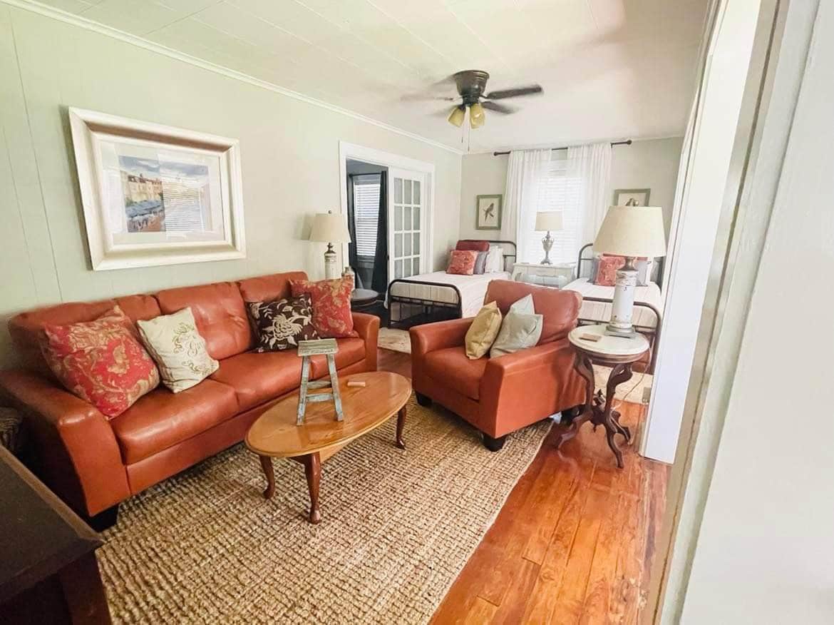 A comfortably furnished living area features a leather sofa and armchair, accompanied by a wooden coffee table. Natural light fills the space through a window, and a decorative rug enhances the floor. A ceiling fan provides airflow, creating a relaxed ambiance.