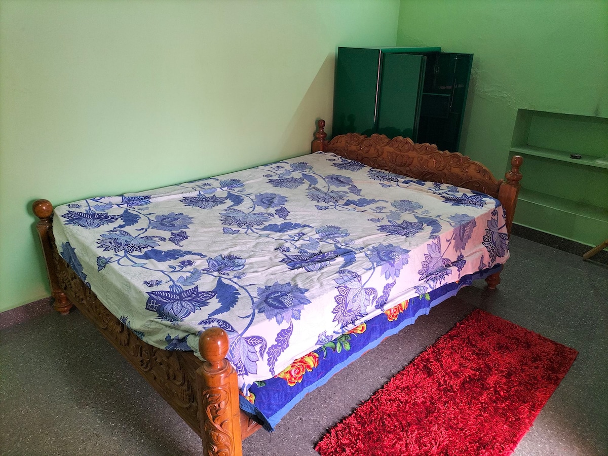 A spacious bed with a carved wooden frame is centered in the room, covered with a floral patterned blanket. A plush red rug is placed at the foot of the bed, while a green cupboard is visible in the background against light green walls.