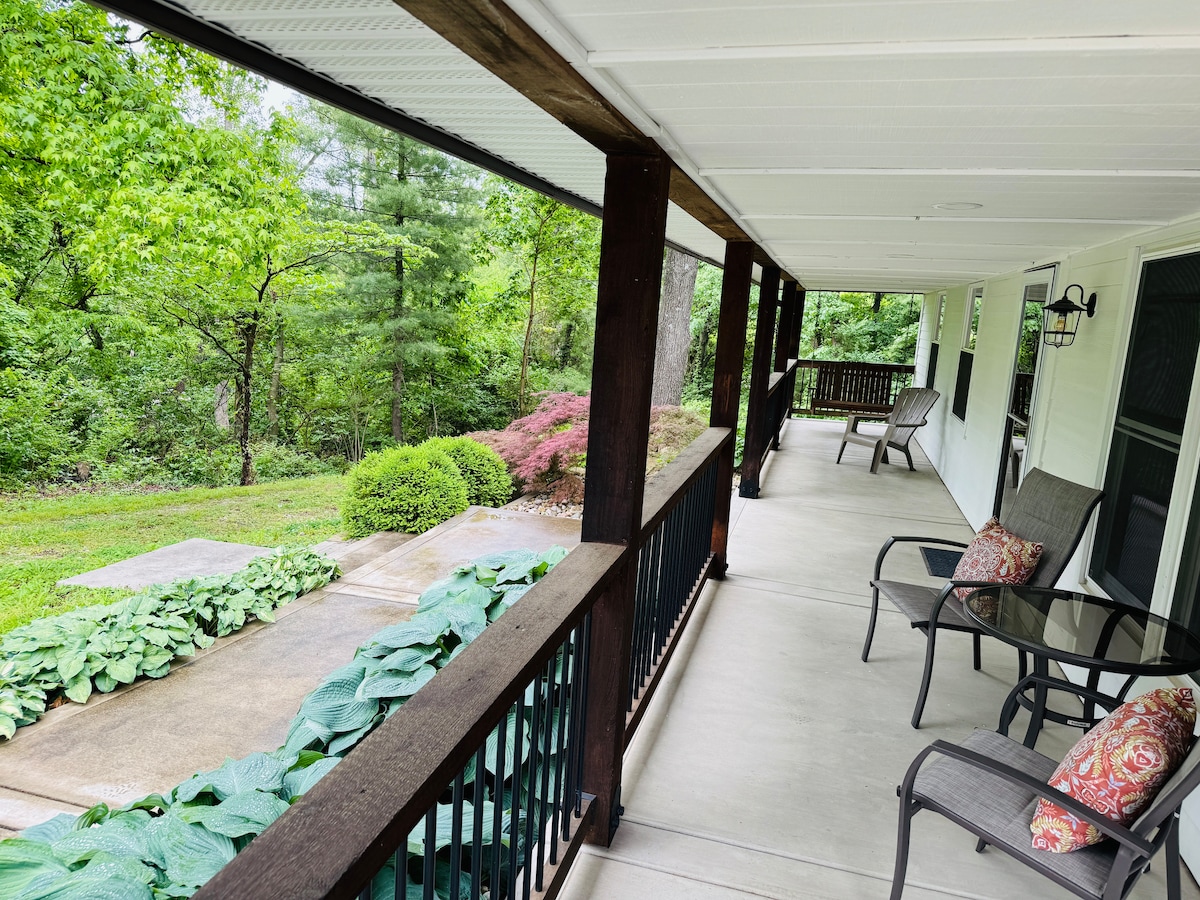 The covered porch features several seating options, including two chairs with cushions. A concrete walkway leads to a lush, green landscape adorned with various plants and trees. The porch offers a view of the surrounding woods, creating a tranquil outdoor space.