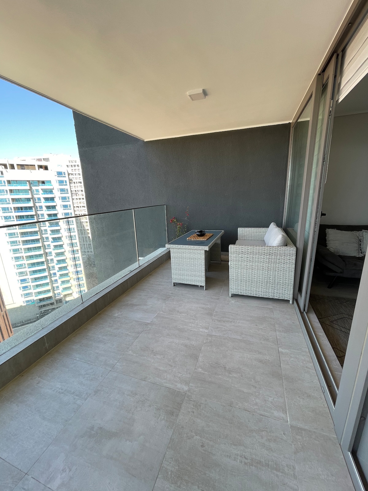 A private balcony features modern outdoor furniture, including a small table and two wicker chairs. The space is lined with glass railings, offering an unobstructed view of the surrounding cityscape. Neutral tile flooring enhances the area, providing a serene setting for relaxation.