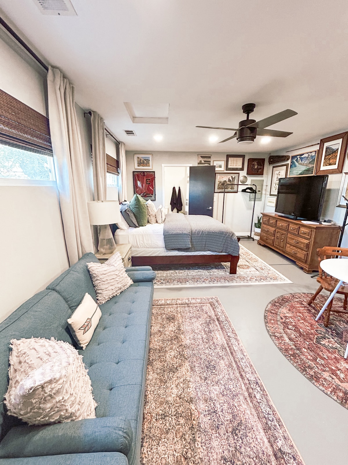 The suite features a spacious sleeping area with a queen bed, complemented by a cozy blue sofa nearby. A television is mounted on a wooden dresser, while a small dining table is set for four. Natural light filters through window treatments.