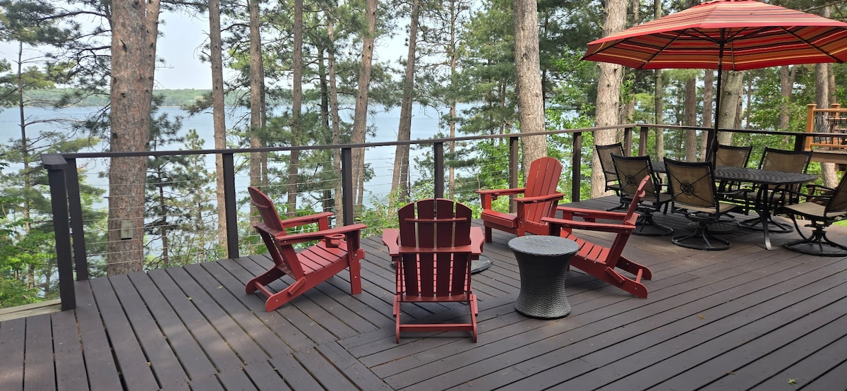 A spacious wooden deck features four red Adirondack chairs arranged around a dark side table, providing a comfortable seating area. An umbrella-shaded table with multiple chairs is also visible, surrounded by trees and offering views of the lake in the background.