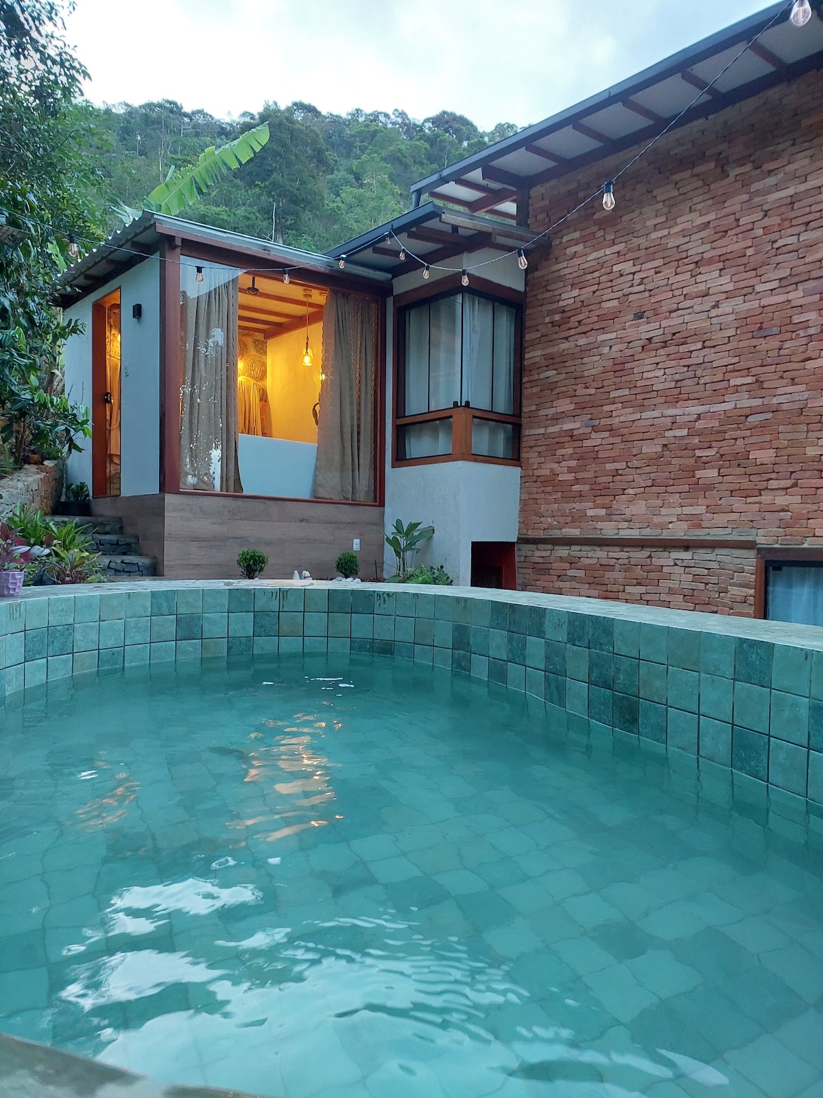 A serene outdoor space features a clear pool, surrounded by light blue tiles. The backdrop includes a rustic stone wall and large windows allowing natural light to enter the interior. Lush greenery is present, enhancing the peaceful atmosphere of the area.