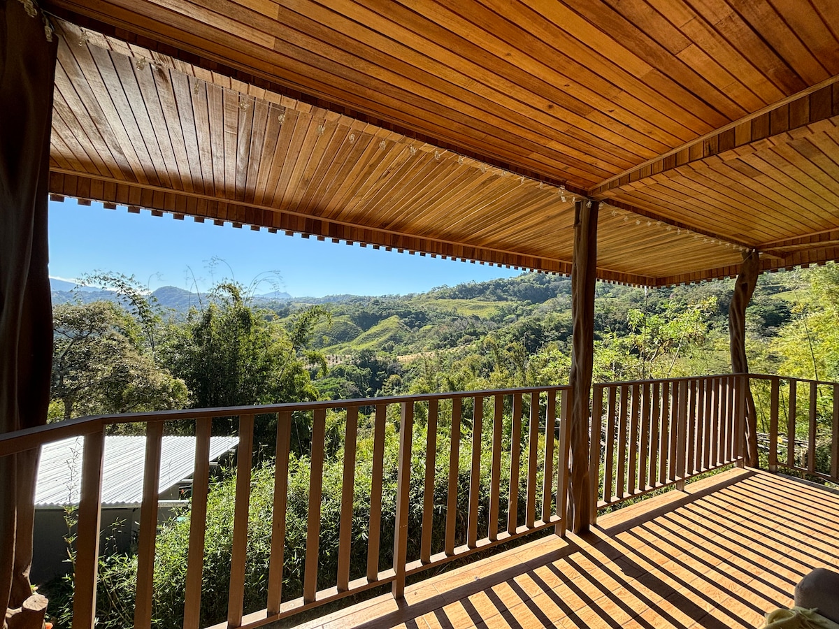 A wooden balcony offers expansive views of the surrounding hills and greenery. The ceiling features elegant wooden planks, and the railing provides a natural border. Sunlight filters through, illuminating the space and enhancing the peaceful setting.