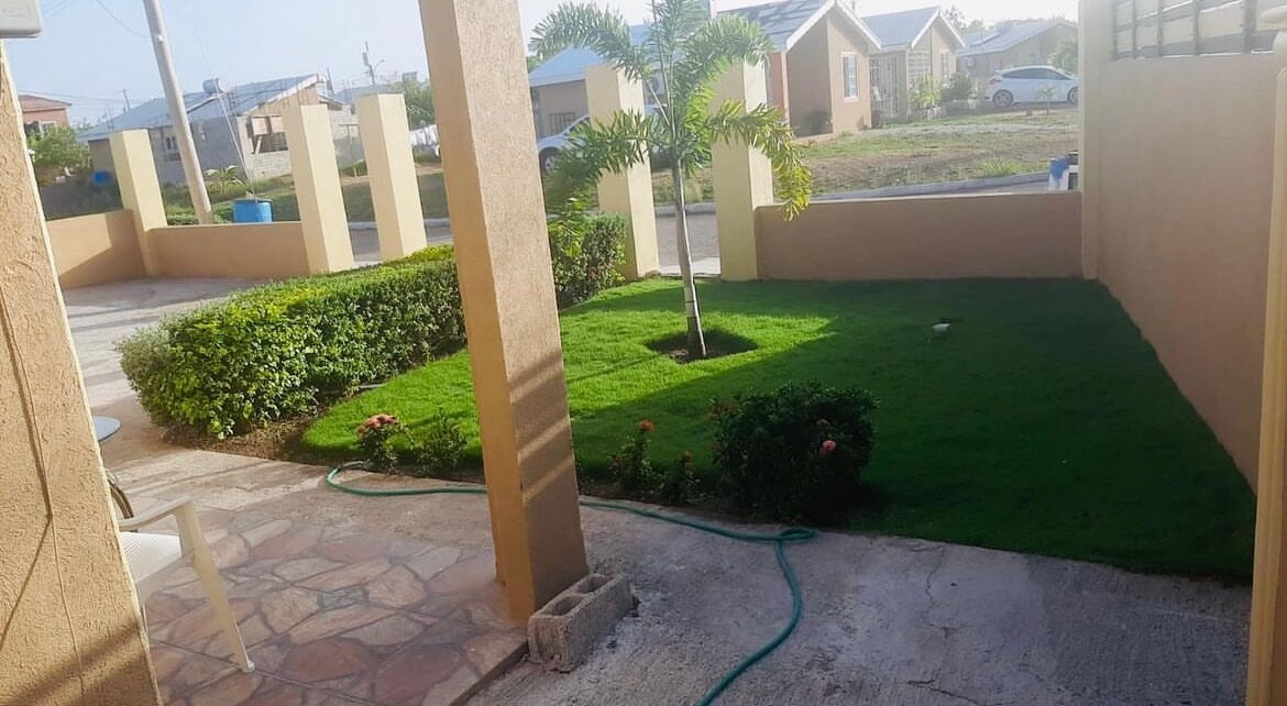 A well-maintained front yard features a small green lawn complemented by neatly trimmed shrubbery and a young palm tree. Paved stones lead towards the entrance, while colorful flowers add a touch of vibrancy to the landscape. The peaceful residential street is visible in the background.