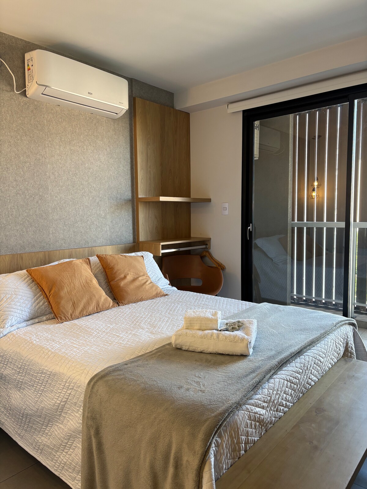 A modern bedroom is displayed, featuring a spacious bed dressed in a light quilt and accent pillows. Shelving is positioned above the bed, and a desk is visible beside it. Bright light filters in through glass doors that open to a balcony with a railing.