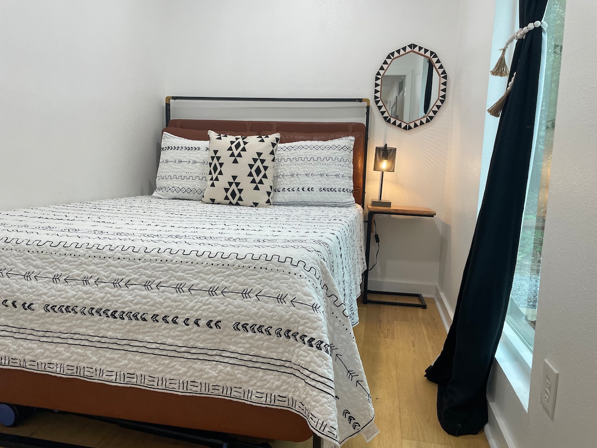 A cozy bedroom features a spacious bed adorned with a patterned blanket and decorative pillows. A bedside table with a lamp offers soft lighting, while a round mirror reflects the room's brightness. Natural light enters through a window draped with dark curtains.
