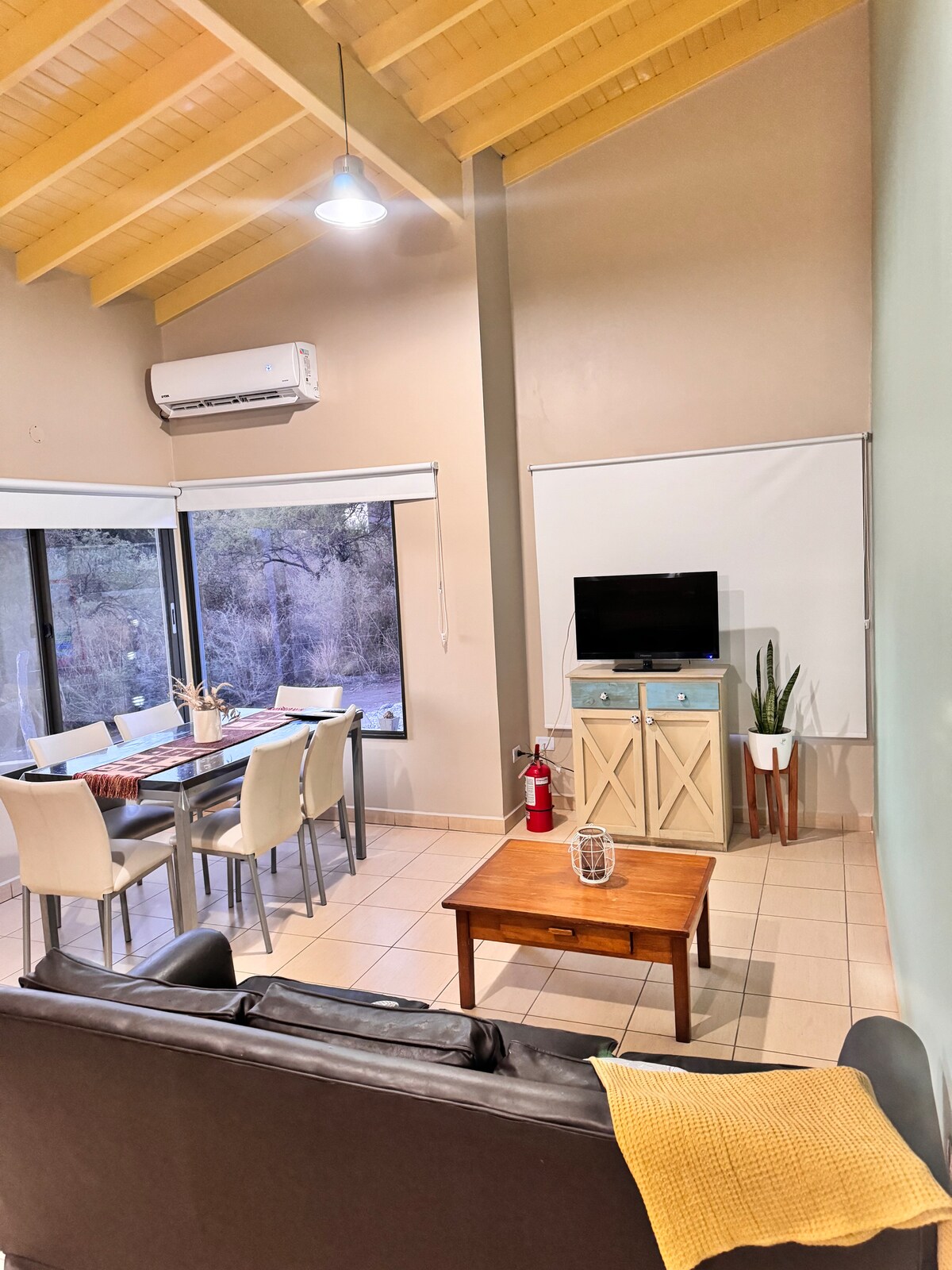 An open living and dining area features high ceilings with wooden beams, large windows allowing natural light, and a comfortable dark sofa. A TV is mounted on the wall, and a dining table with light-colored chairs is set for meals, enhancing the functional space.