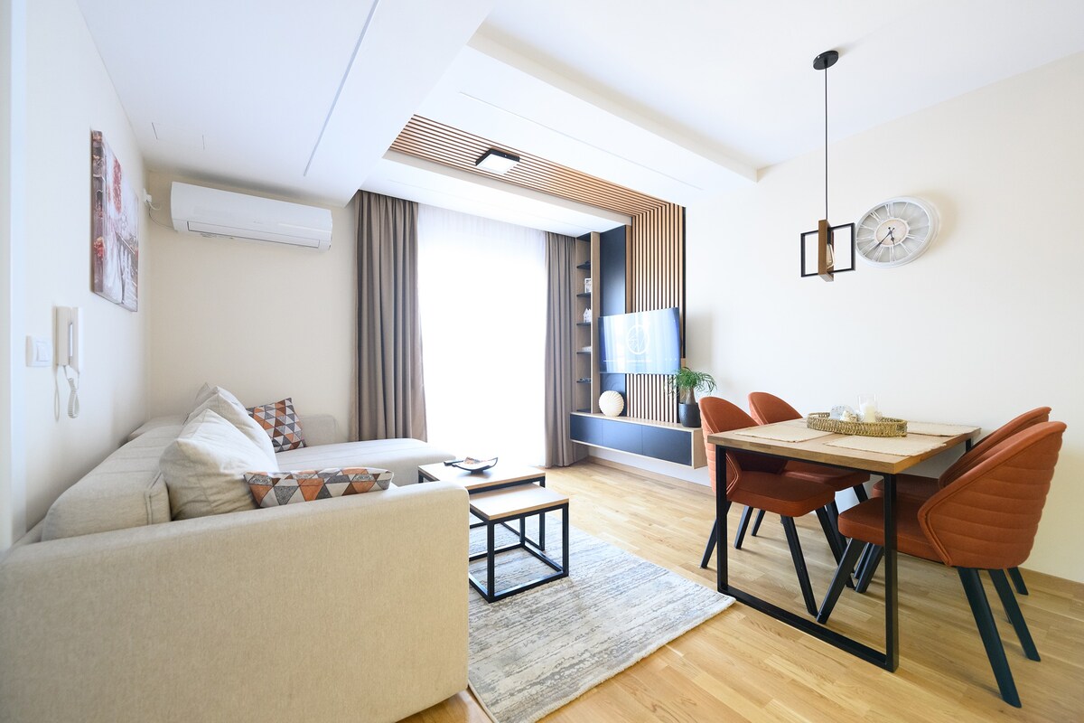 A modern living area features a light-colored sofa with patterned cushions, paired with a glass coffee table. A dining table with four orange chairs sits adjacent, and a large window allows natural light to fill the space. Decorative elements add a contemporary touch.