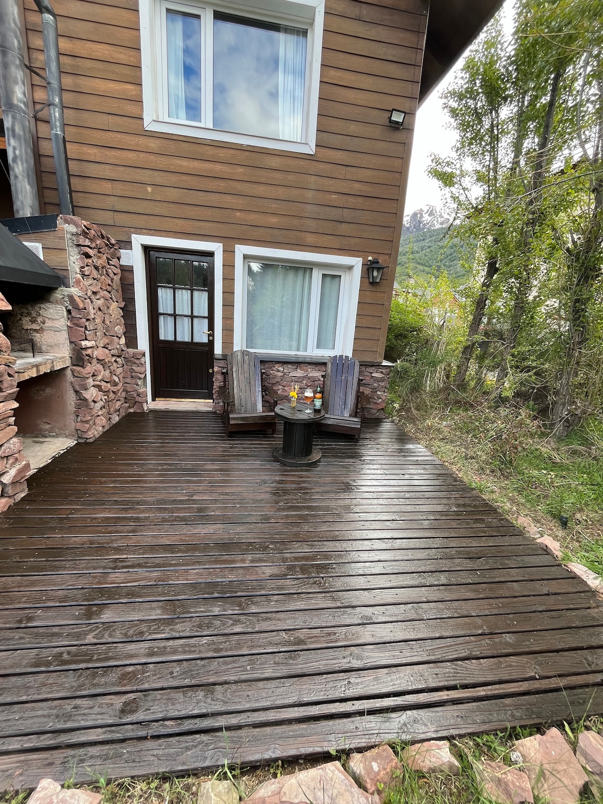 A wooden deck area is seen, featuring two dark wooden chairs positioned around a small table. The entrance to the interior is visible, along with a stone wall that adds texture. Lush greenery surrounds the space, creating a serene outdoor setting.