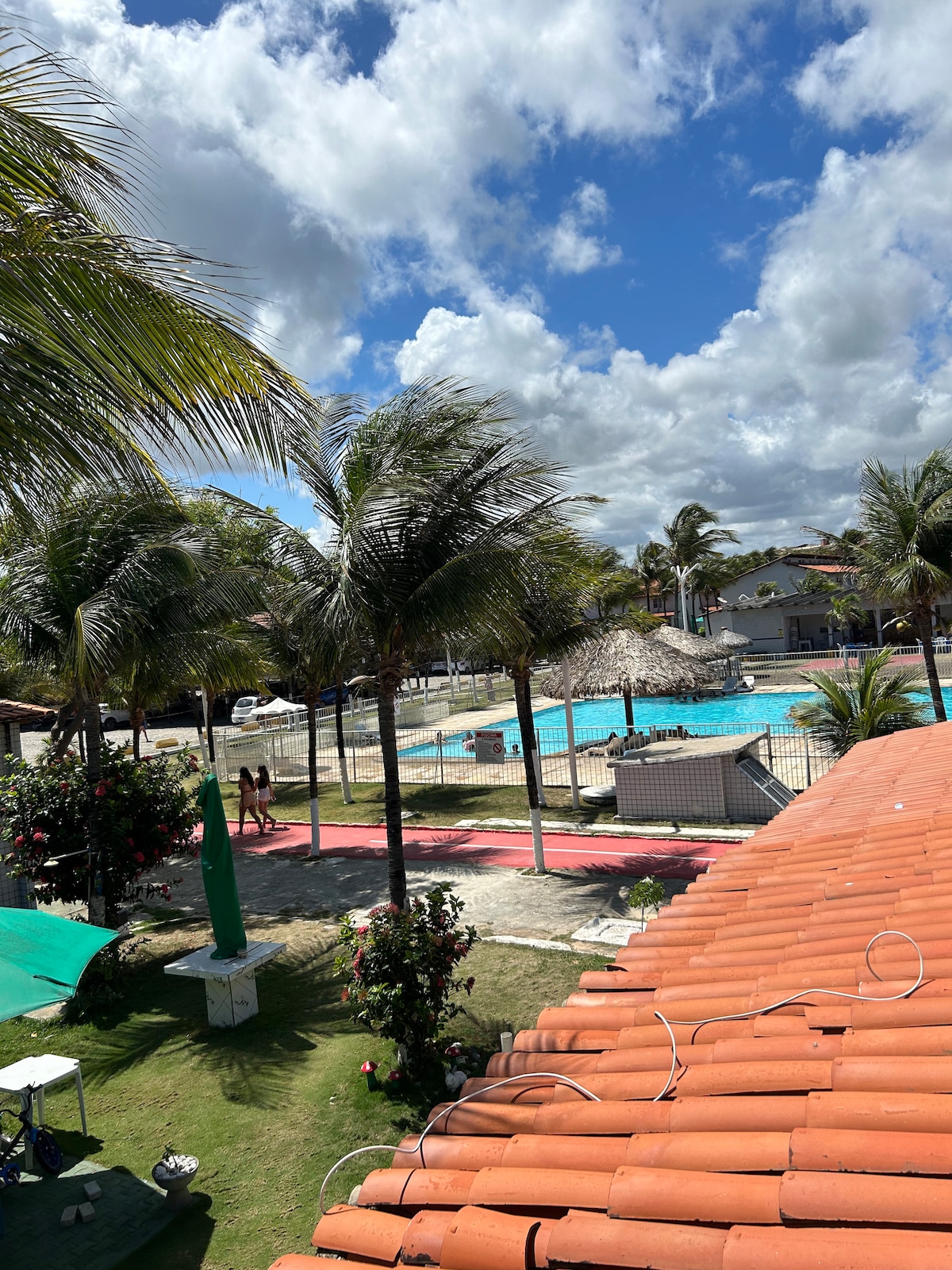 A view of an outdoor area featuring a blue swimming pool surrounded by palm trees. A poolside gazebo is visible alongside a grassy space with pathways. The red-tiled roof of a nearby building adds a contrasting color to the scenery, enhanced by scattered clouds in a bright sky.