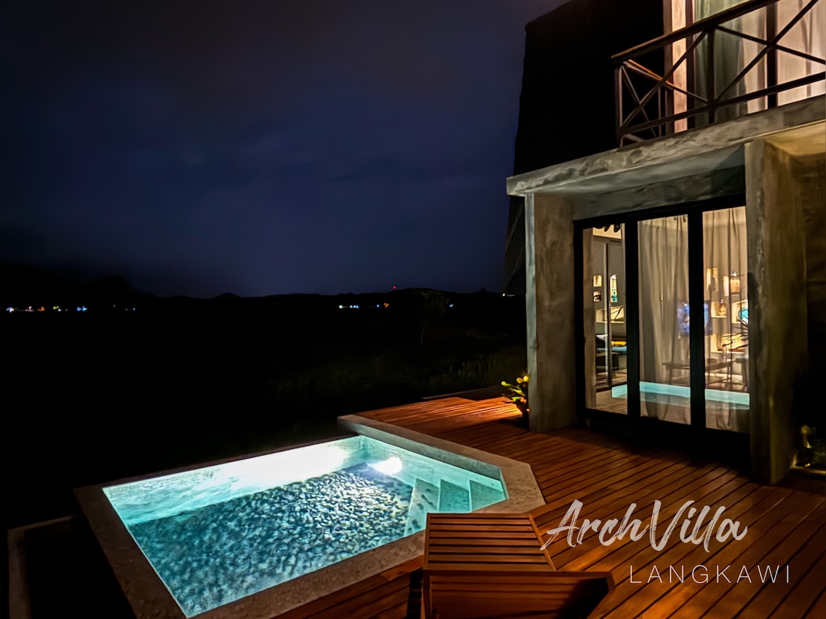 The villa's exterior is illuminated at night, showcasing a private swimming pool with soft blue lighting. The wooden deck extends from the living area, creating a seamless connection between indoor and outdoor spaces. The serene landscape features distant lights twinkling across the horizon.