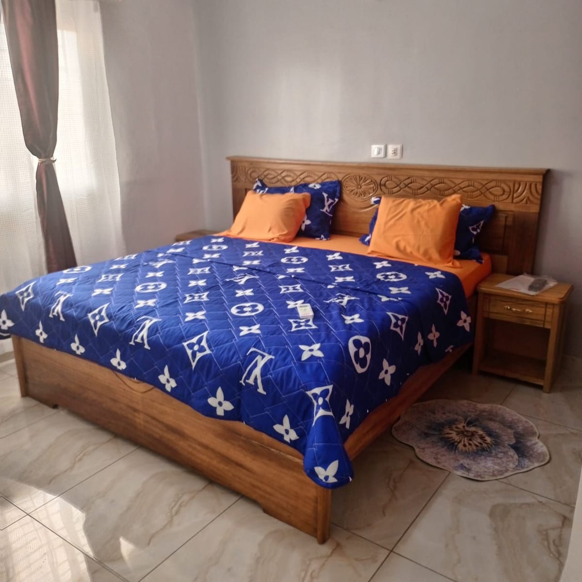 A spacious bed is adorned with a vibrant blue and orange patterned blanket. Two matching pillows complement the colorful bedding. A bedside table is positioned next to the bed, contributing to the room's modern design. Soft light filters through a nearby window, enhancing the atmosphere.