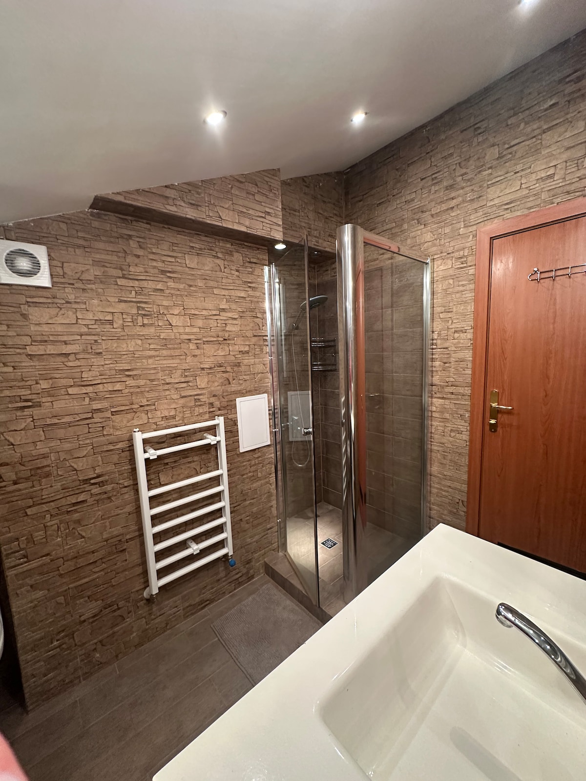 A modern bathroom features a sleek glass shower enclosure and a spacious white bathtub. The stone wall adds a natural touch, while a heated towel rack is mounted nearby. Warm lighting is provided by recessed ceiling fixtures, enhancing the ambiance.