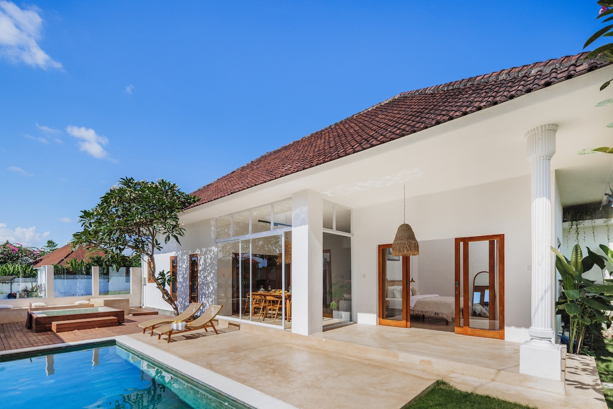 The villa's exterior showcases modern architecture with large glass doors that connect the indoor and outdoor areas. A spacious terrace is framed by tropical plants, featuring a private swimming pool and sun loungers, all set against a clear blue sky.