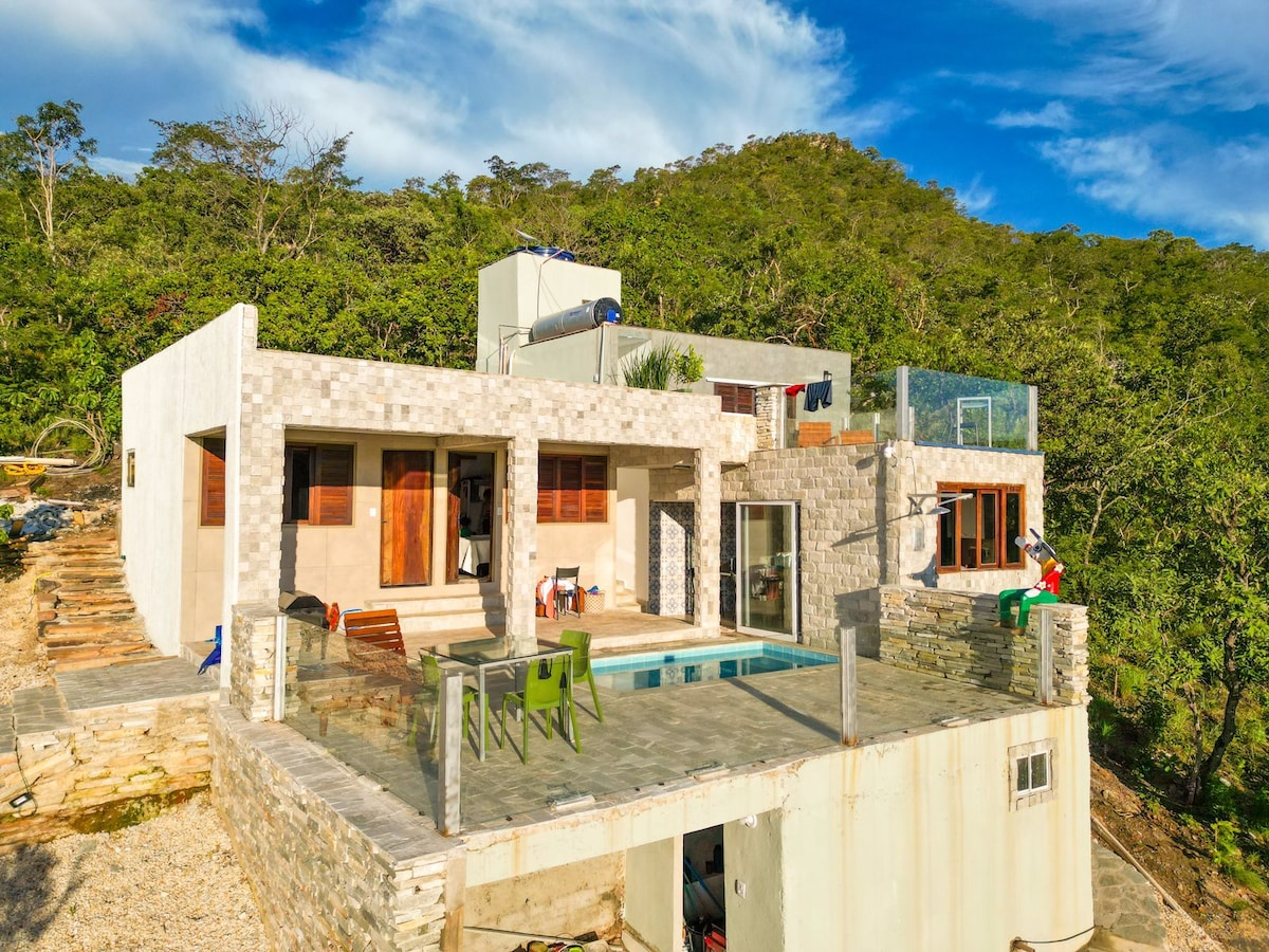 The exterior of a modern villa showcases a combination of stone and concrete textures against a lush, green hillside. A spacious deck features a private pool. Large glass doors invite natural light into the interior, where seating areas and outdoor furniture are positioned.
