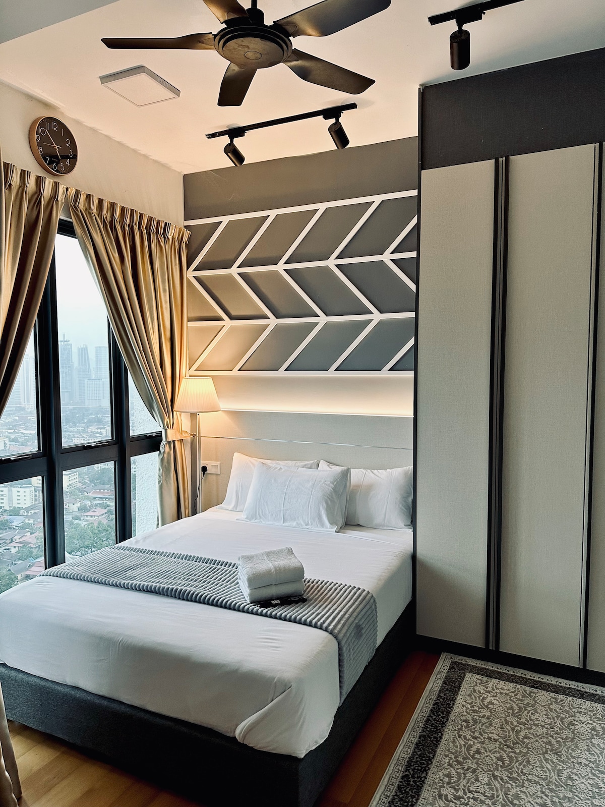 A cozy bedroom is arranged with a spacious bed adorned with soft linens and pillows. Decorative elements include a sleek geometric accent wall and a lamp providing warm light. Large windows offer a view of the city, framed by elegant curtains.