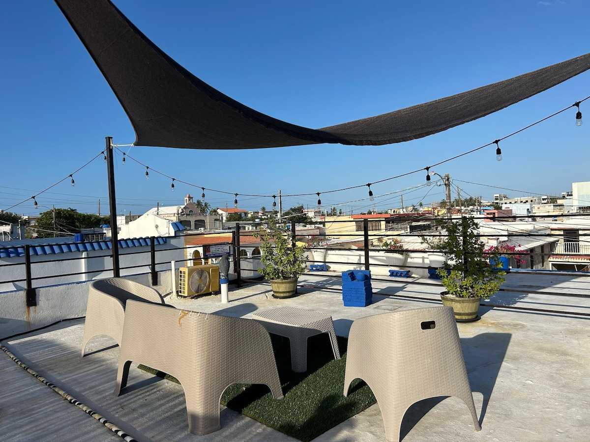 A rooftop terrace features a shaded area under a dark canopy, providing a comfortable space with several light-colored seating arrangements. Potted plants add greenery, while string lights are suspended overhead. The urban landscape of rooftops is visible in the background, under a clear blue sky.