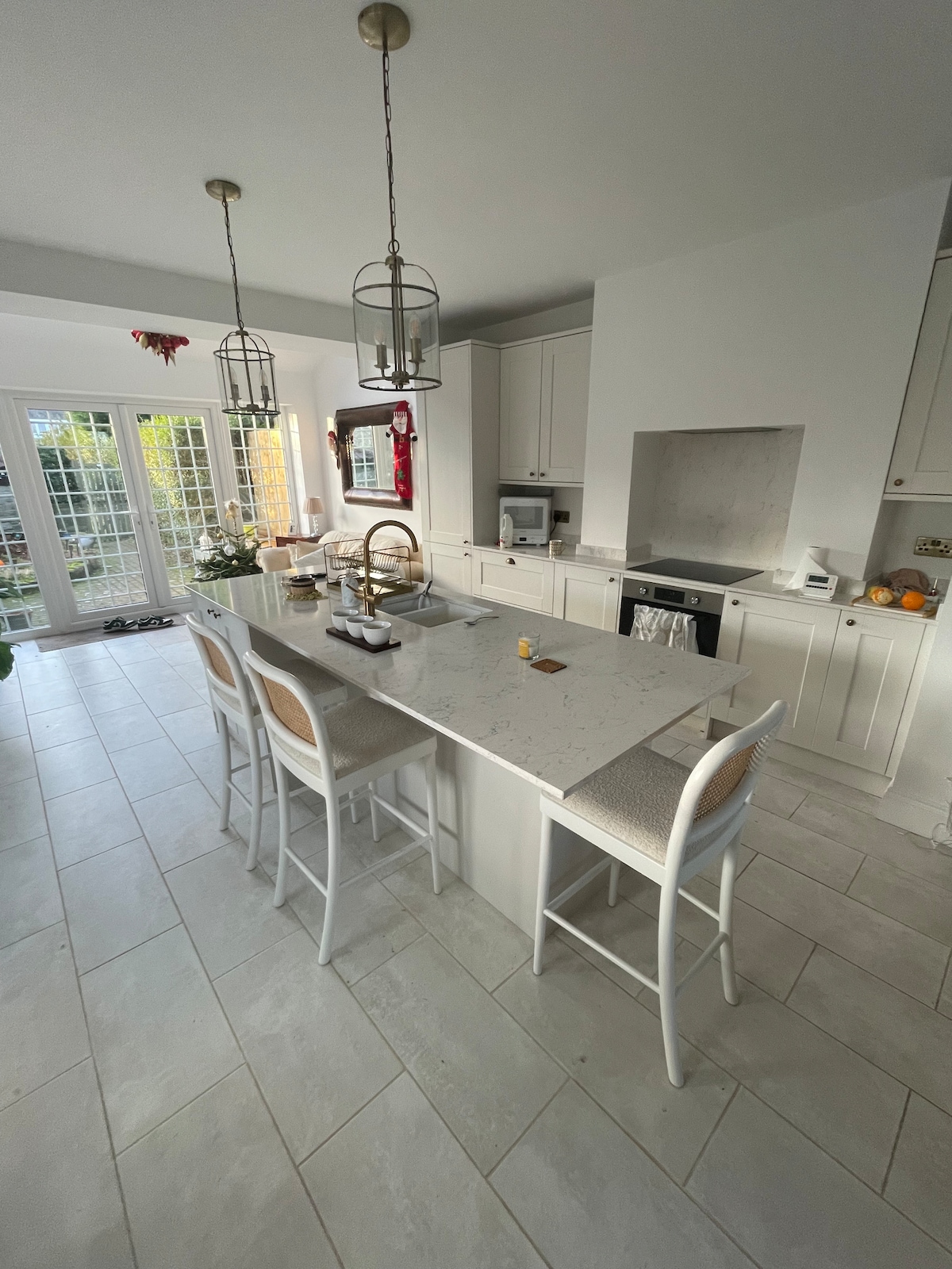 A spacious kitchen area features a long island with four bar stools, surrounded by modern cabinetry. Light-colored tiles cover the floor, and natural light streams through large glass doors leading to an outdoor space. Minimal decorative elements contribute to the clean aesthetic.