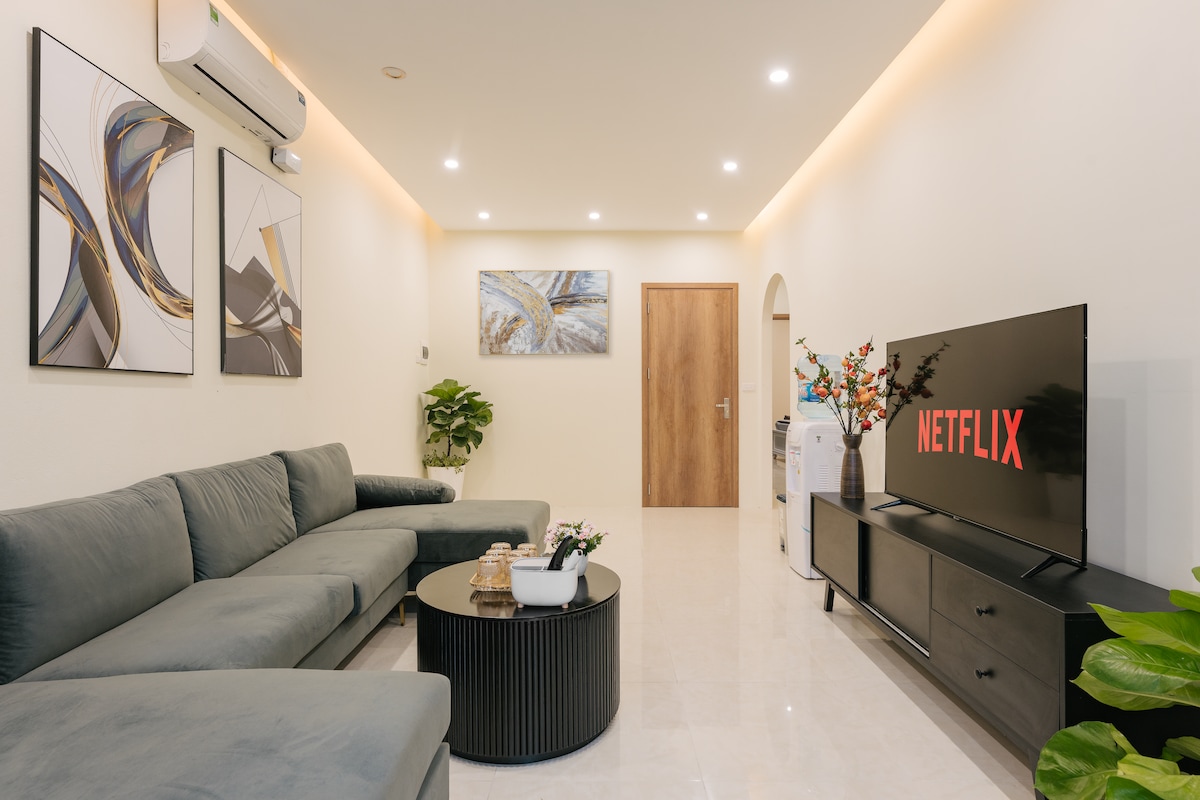 A modern living area features a large gray sectional sofa arranged around a round black coffee table. A flat-screen television displaying the Netflix logo is positioned on a low black cabinet. Light artwork adorns the walls, complemented by indoor plants, adding a relaxed ambiance.