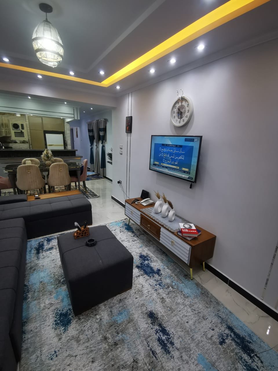 A modern living area showcases a spacious sectional sofa with a sleek coffee table in front. A television is mounted on the wall, complemented by decorative items on a low cabinet. Soft lighting is provided by an elegant ceiling fixture, enhancing the comfortable atmosphere.