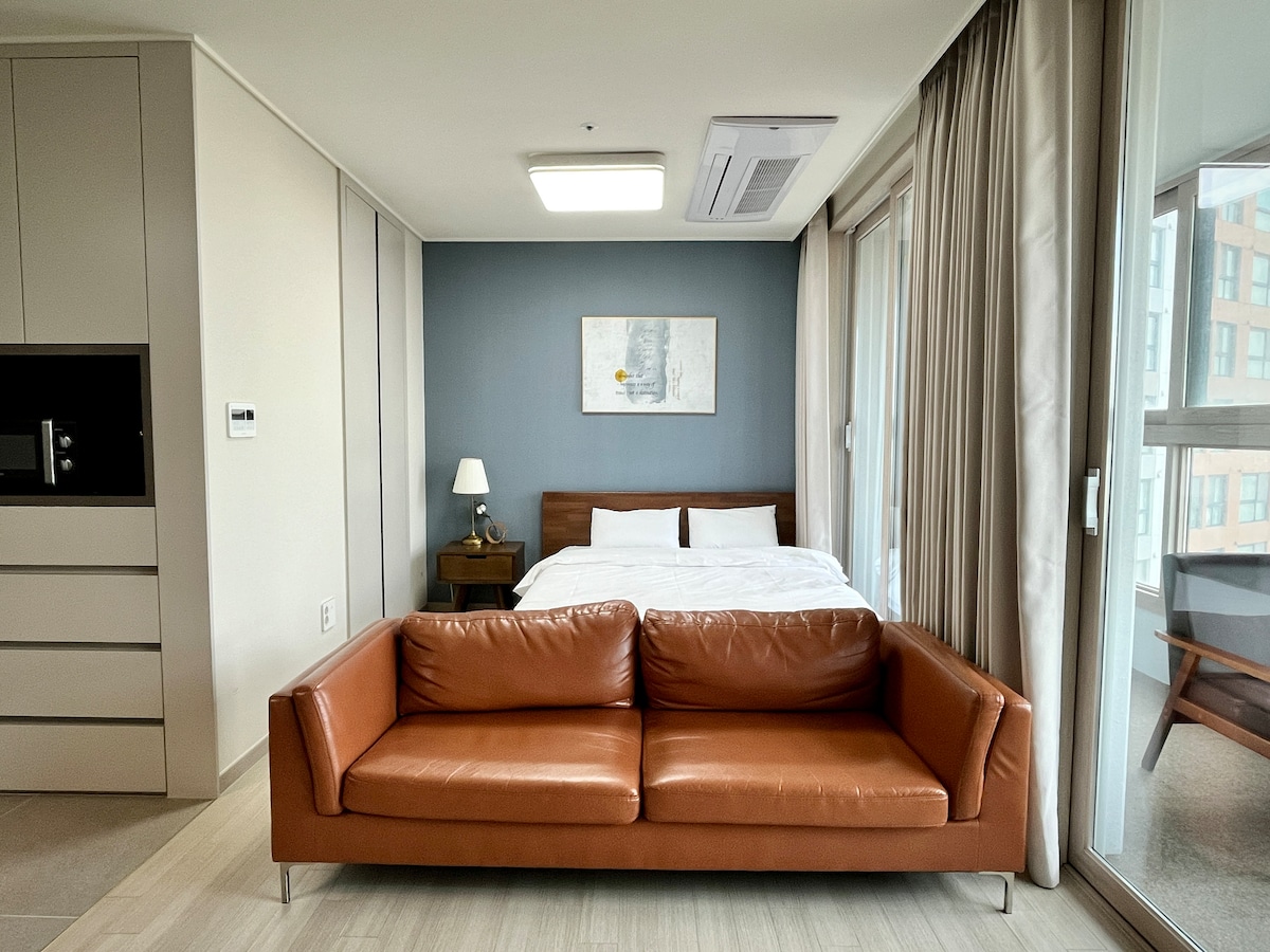 A modern living area features a brown leather sofa facing a bright sleeping area. Natural light filters through large windows, which are adorned with soft curtains. In the background, a neatly made bed is complemented by minimalistic decor and a small bedside lamp.