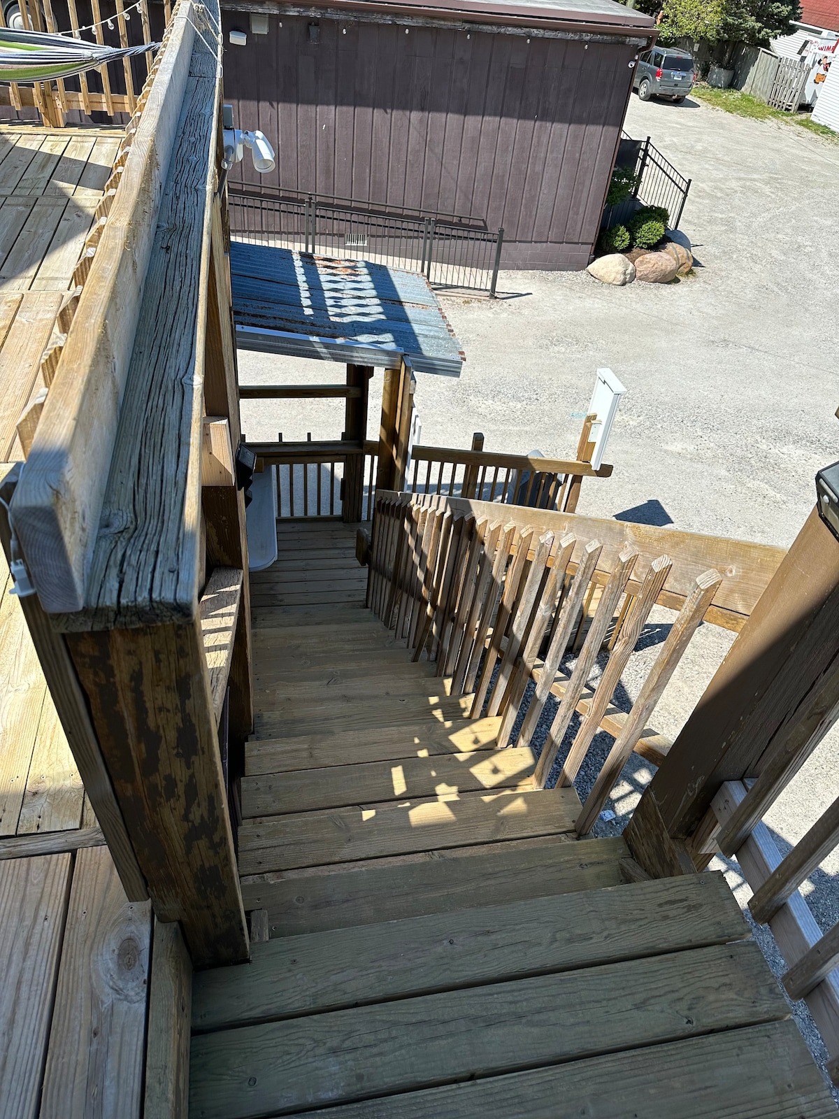 A wooden staircase with parallel railings is seen descending from a patio area. Sunlight casts shadows on the steps, highlighting the natural wood grain. The surrounding area includes a gravel parking lot and nearby buildings.