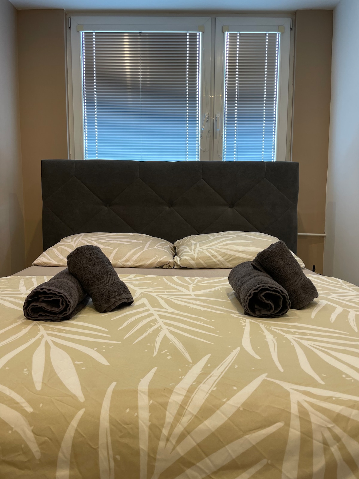 A cozy sleeping area is presented with a patterned duvet cover featuring a leafy design. Two neatly rolled towels are positioned on each side of the spacious bed, which is framed by a grey upholstered headboard. Natural light enters through the window covered with blinds.