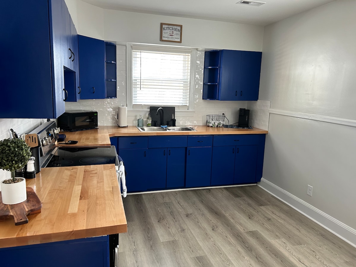 A well-equipped kitchen features deep blue cabinetry and warm wooden countertops. The space includes modern appliances, a sink, and a window allowing natural light to enhance the room. Shelving and organizational elements provide ample storage for kitchen essentials.