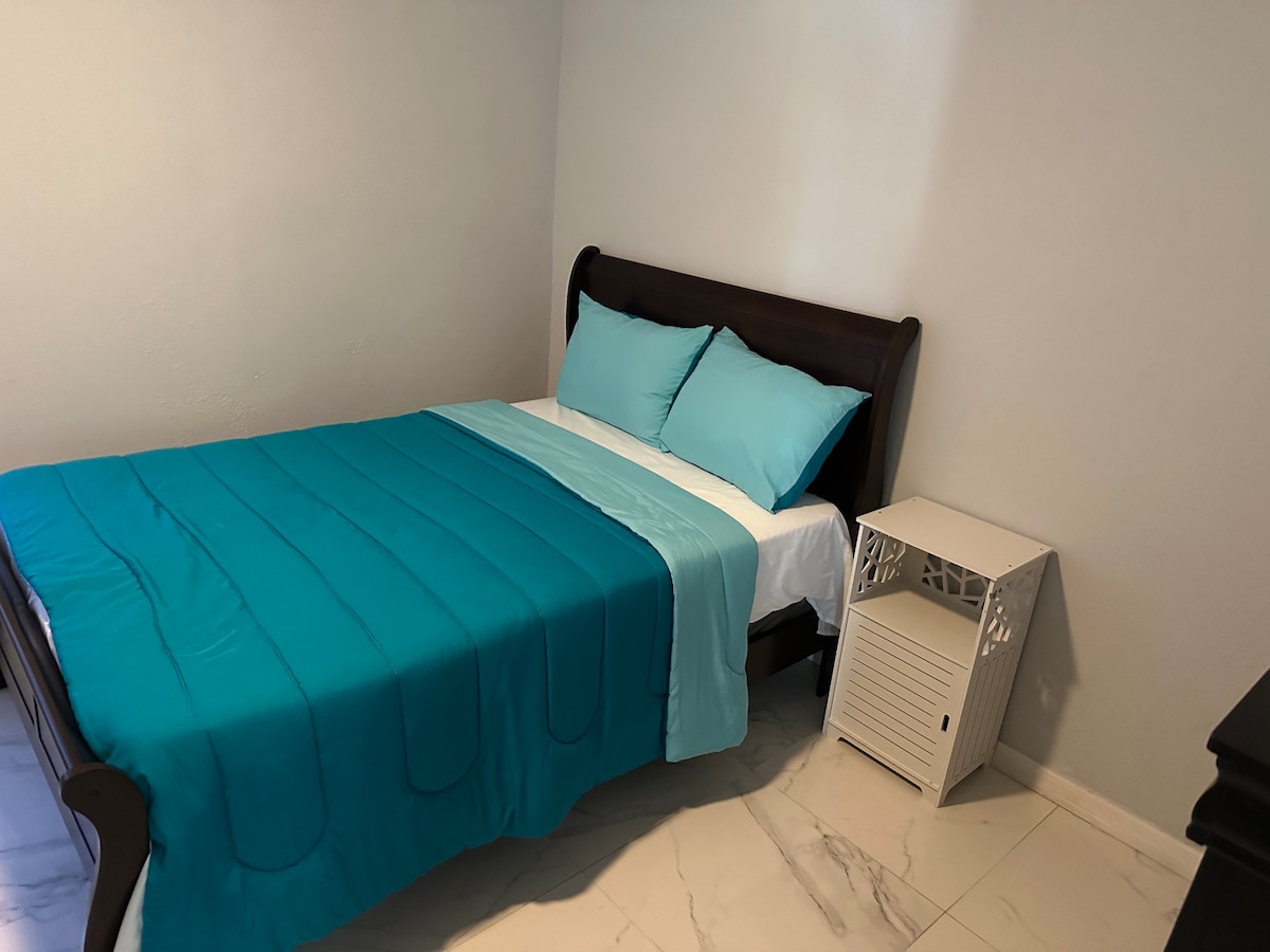 A cozy bedroom features a wooden bed frame with a spacious mattress dressed in a vibrant turquoise coverlet. Two matching pillows rest against a light-colored headboard. A small white nightstand is positioned beside the bed, complementing the light-colored walls and marble flooring.