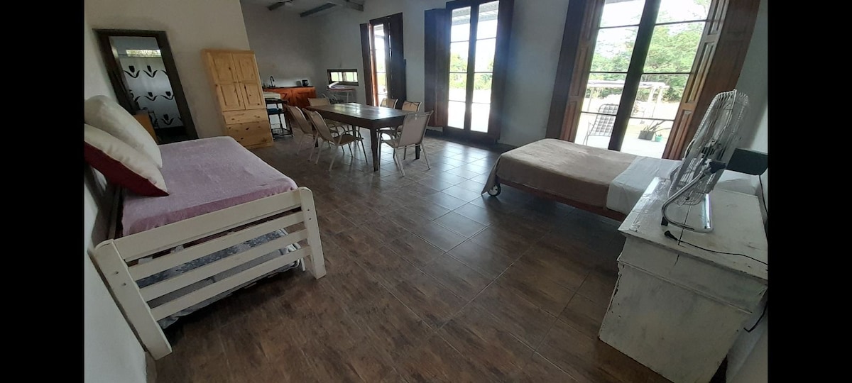 An open living space is presented, featuring a combination of seating and sleeping areas. Two beds are positioned alongside a dining table surrounded by chairs. Large windows allow natural light to fill the room, with views of greenery visible outside.