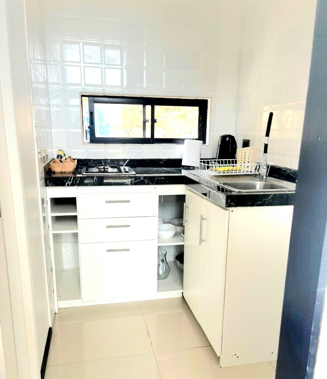 A compact kitchen area is presented with sleek white cabinetry and a dark countertop. Essential appliances, including a kettle and a stovetop, are visible. Natural light enters through a small window, illuminating the organized space featuring dishware and cooking essentials.