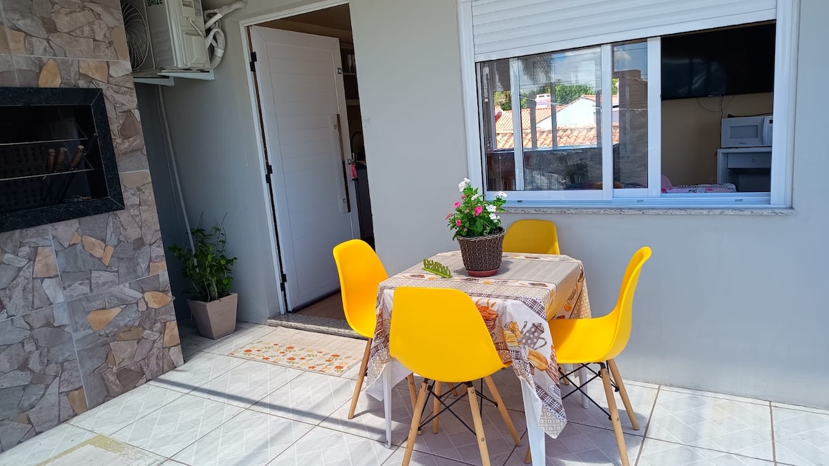 A small dining area is set on a tiled patio, featuring a round table with a floral tablecloth. Four yellow chairs surround the table, and a potted plant adds a touch of greenery. A stone grill is visible nearby, creating an inviting outdoor space.