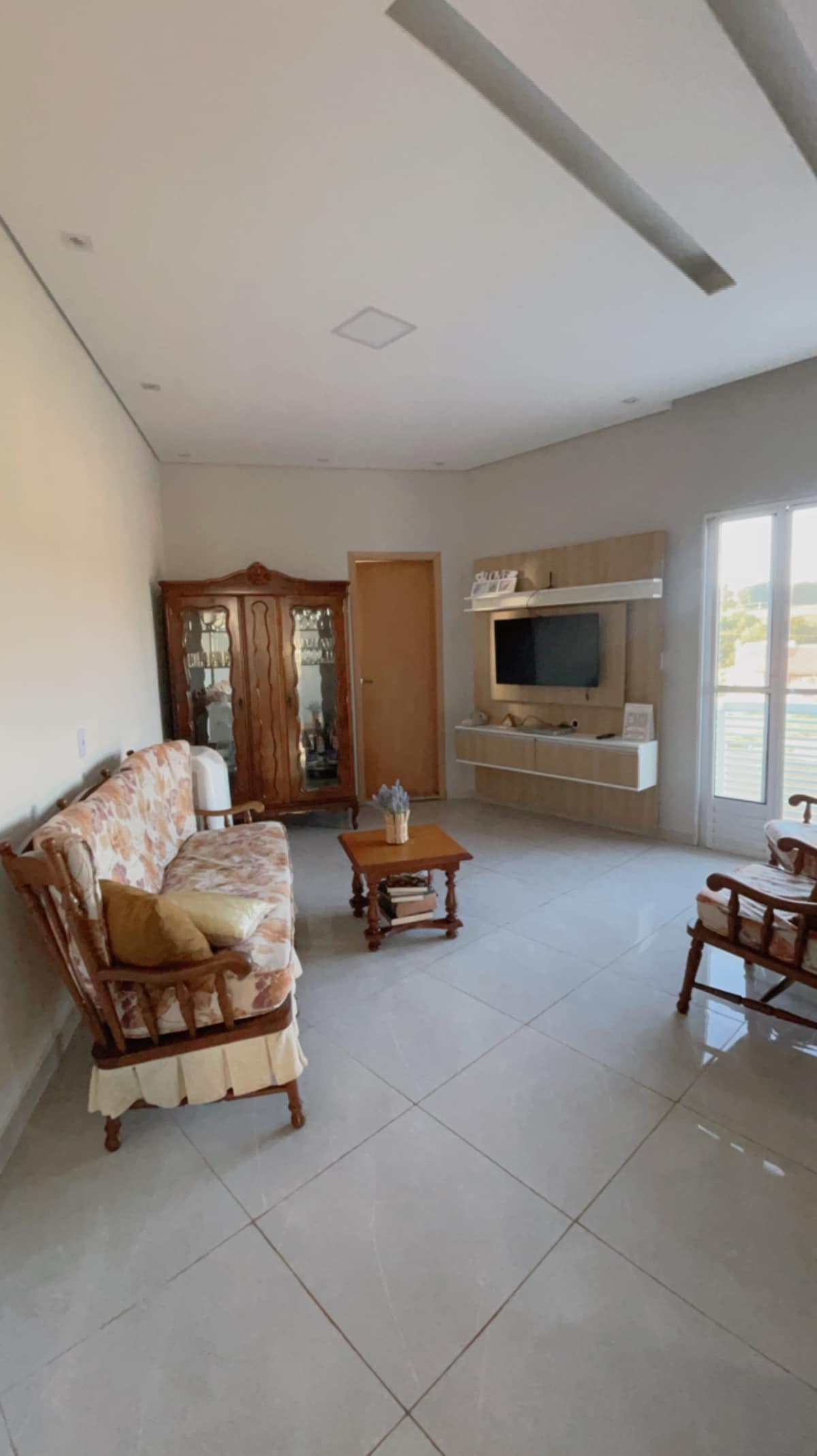A living area showcases a light-colored sofa with floral patterns across from a wooden TV stand. A glass cabinet displays decorative items, while two rocking chairs provide additional seating. Natural light fills the space through large doors leading to a balcony.