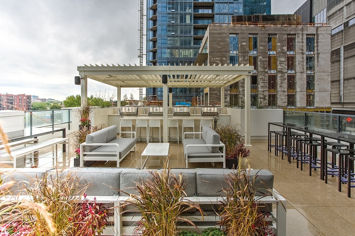 An outdoor terrace is featured with multiple seating areas, including gray sofas and high-top tables. Lush plants surround the space, and a canopy offers partial shade. The backdrop showcases urban buildings, contributing to a contemporary setting.