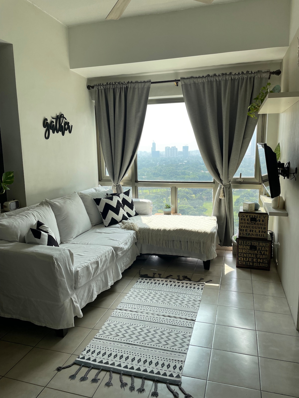 A comfortable seating area is arranged near large windows, offering a view of the green golf course and cityscape. Neutral-toned curtains frame the windows, and a cozy area rug is placed on the tiled floor. A small television is mounted on the wall, complemented by decorative storage boxes.