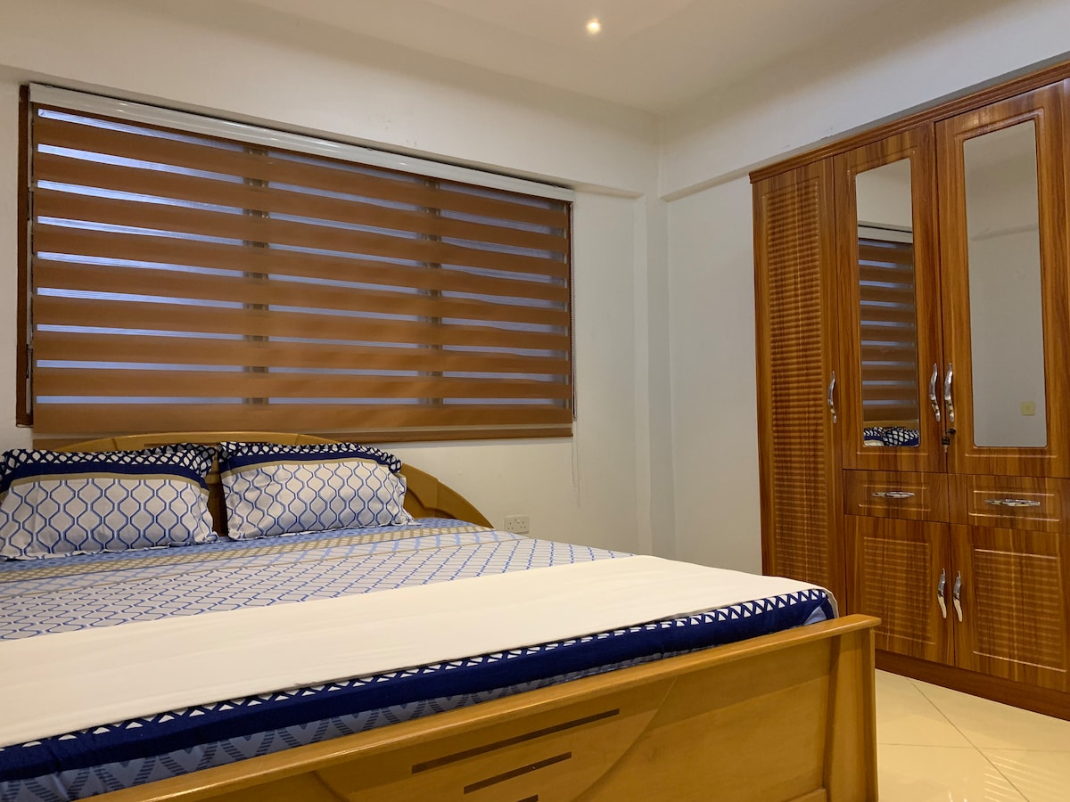 A spacious bedroom features a large wooden bed with a blue-patterned duvet cover and coordinating pillows. A wooden wardrobe with mirrored doors is positioned against the wall, while light filters through the stylish horizontal window blinds.