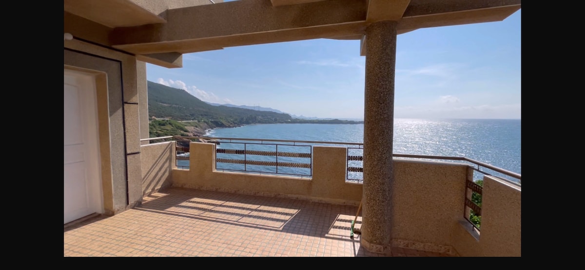 A spacious terrace is shown, offering expansive views of the sea and coastline. The golden sunlight reflects off the water, creating a shimmering effect. The area features railings and textured flooring, providing a pleasant outdoor space for relaxation.